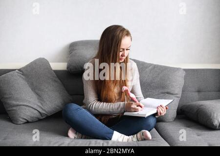 Creative pensive young female sitting on the couch at home writing something in her journal. Full body portrait. Distance learning or working concept. Stock Photo