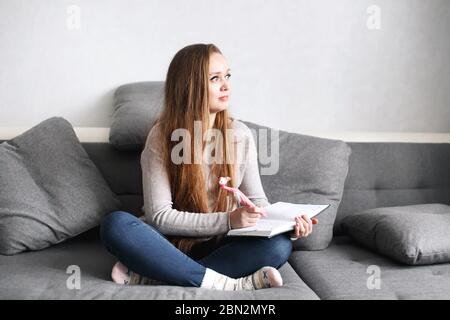 Creative pensive young female sitting on the couch at home writing something in her journal. Full body portrait. Distance learning or working concept. Stock Photo