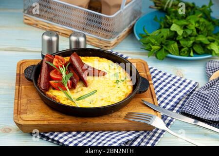 Omelet with sausage and tomatoes in a pan on the table. Appetizing breakfast. A sprig of rosemary, a wooden spice. Hunt sausages. Tasty food fried Stock Photo