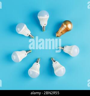 A Golden light bulb stands out in the circle of light bulbs of a ring of white lamps on a blue background. One original idea out of many is mediocre. Stock Photo