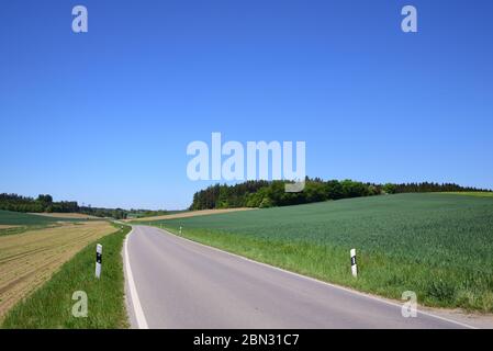 A narrow country road winds through romantic green Bavarian countryside, with blue skies, in spring Stock Photo