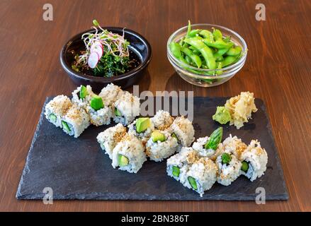 Japanese sushi food with avocado, rice, sea delicacies on a black stone, on a brown background. Plates with salad and green beans. Stock Photo