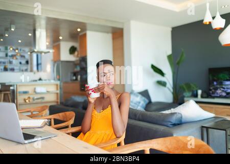 Thoughtful young woman drinking coffee, working from home at laptop in dining room Stock Photo