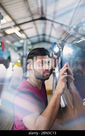 Focused male mechanic with flashlight examining scratches on car in auto repair shop Stock Photo