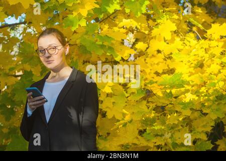 On a warm autumn day, a woman is looking slightly surprised at the camera while holding her smartphone at chest level. Tranquil outdoor environment. Stock Photo