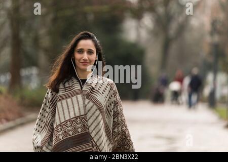 Young Indian woman wearing poncho and earphones smiling for the camera outdoors. Medium shot. Stock Photo