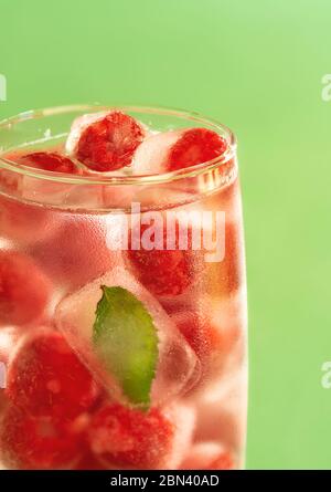 Frozen raspberries and mint leaf in a glass of water on a green background. Glass of water with backlight. Stock Photo