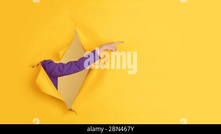 Pointing. Female hand gesturing in torn yellow paper hole background. Breaking on, breakthrought. Concept of business, finance, shopping, proposal, sales, ad. Copyspace Promotions discounts Stock Photo