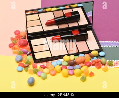 Red, scarlet lipstick and palette with mirror lay on peachy and yellow background near dragee sweets and candied fruit. Make up concept. Beauty supplies, lipstick, palette, powder, rouge, concealer. Stock Photo