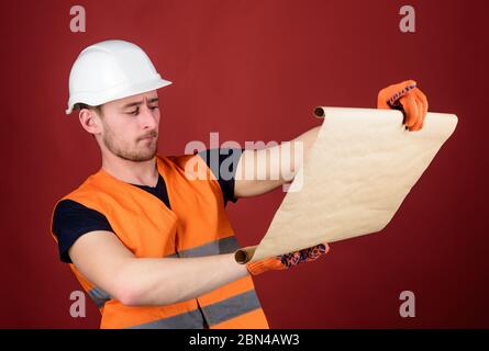 Foreman concept. Engineer, architect, builder on strict face holds blueprint in hands, supervises construction site. Male contractor with house plans wearing hard hat, controls works, red background Stock Photo