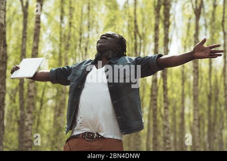 Happy African man with notebook and headphones with outstretched hands enjoying the freedom surrounded by trees Stock Photo