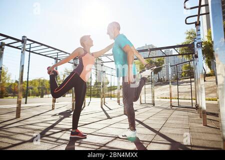 Happy mature man and woman in sportswear warming up together at the stadium in the morning. They stretching legs, looking at each other and smiling Stock Photo