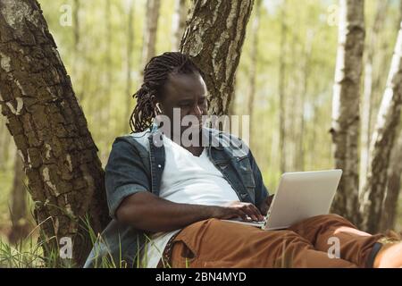 Cheerful african man doing remote work on the laptop in the nature.  Stock Photo