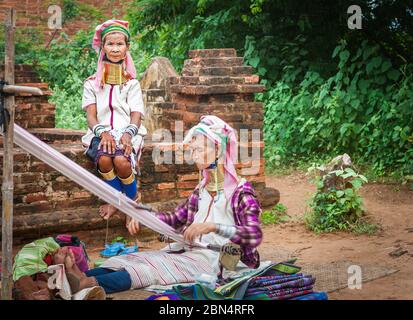 Bagan Myanmar - October 29 2013; Two Kayan women with brass rings around necks one weaving while other in selective focus sitting wall looks on. Stock Photo