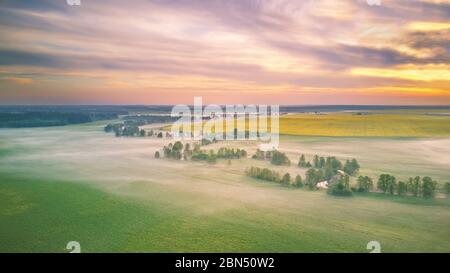 Spring morning aerial rural panorama. Sunrise over green and yellow blooming colza fields. Clouds of fog and small river with trees on riverbank. Bela Stock Photo