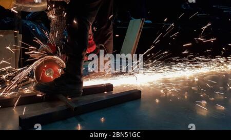 Factory worker in a protective mask using an electric meat grinder tool cuts metal in the dark, sparks fly beautifully apart. Stock Photo
