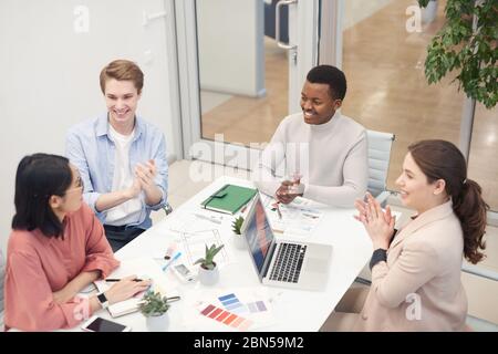 High angle view at multi-ethnic group of business people clapping to Asian colleague while collaborating on design project during meeting in office, c Stock Photo