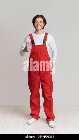 Smiling young laborer posing in work overalls and showing thumb up Stock Photo