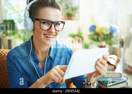 Portrait of happy stylish middle age housewife in jeans shirt with white headphones and tablet PC study online at modern home in sunny day. Stock Photo