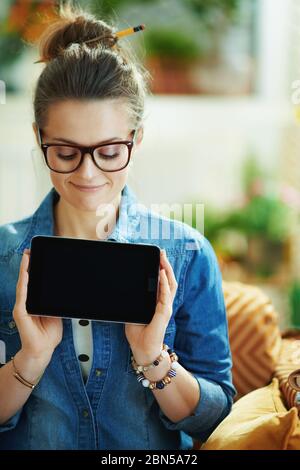 smiling young female in jeans shirt in the modern house in sunny day showing tablet PC blank screen. Stock Photo