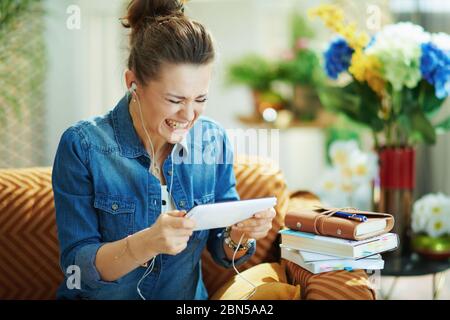 angry young woman in jeans shirt with white headphones and tablet PC study online in the modern house in sunny day. Stock Photo