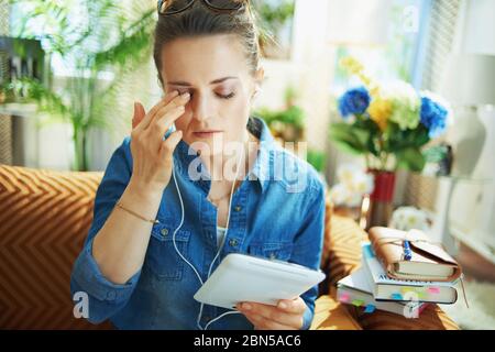 stressed stylish middle age woman in jeans shirt with white headphones and tablet PC study online in the modern living room in sunny day. Stock Photo
