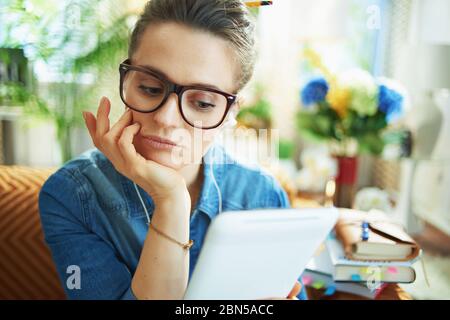 bored modern middle age housewife in jeans shirt with white headphones and tablet PC study online at modern home in sunny day. Stock Photo