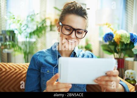 happy modern middle age housewife in jeans shirt with white headphones and tablet PC study online at modern home in sunny day. Stock Photo