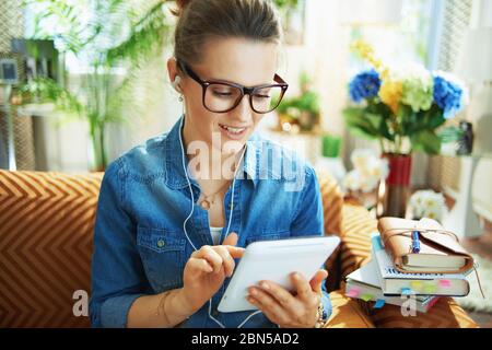 happy modern middle age woman in jeans shirt with white headphones and tablet PC study online in the modern house in sunny day. Stock Photo