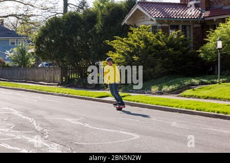A man in a yellow shirt rides an electric one-wheeled skateboard through a residential neighborhood in Fort Wayne, Indiana, USA. Stock Photo