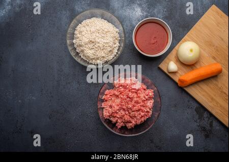 Ingredients for meatballs - minced meat, rice, tomato paste, onion, garlic, carrots on a cutting board on a dark background. Top view Stock Photo