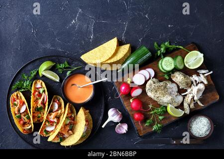 Buffalo Chicken Corn Taco shells loaded with fresh greens and vegetables on a black plate with lime and buffalo sauce with ingredients on a concrete t Stock Photo