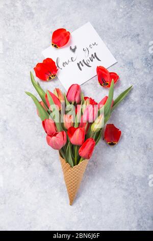 Ice cream of red  tulip flowers in waffle cone with card Love you mom  on concrete  table top view in flat lay style. Stock Photo