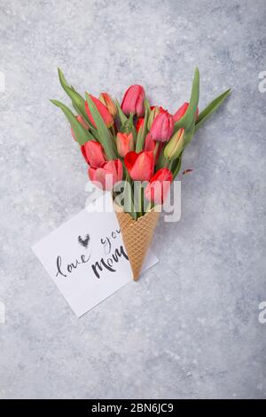 Ice cream of red  tulip flowers in waffle cone with card Love you mom  on concrete  table top view in flat lay style. Stock Photo