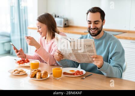 Portrait of young joyful couple using cellphone and reading newspaper while having breakfast in cozy kitchen at home Stock Photo