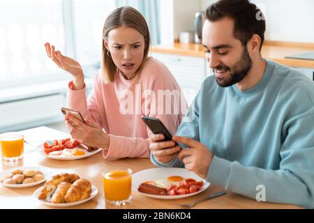 Portrait of young caucasian couple using cellphones while having breakfast in cozy kitchen at home Stock Photo