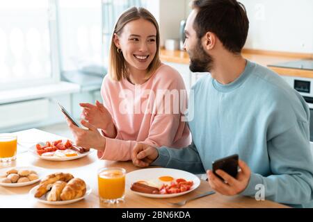 Portrait of young joyful couple using cellphones and laughing while having breakfast in cozy kitchen at home Stock Photo
