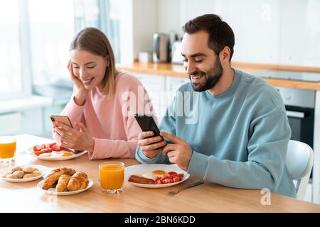 Portrait of young joyful couple using cellphones and laughing while having breakfast in cozy kitchen at home Stock Photo
