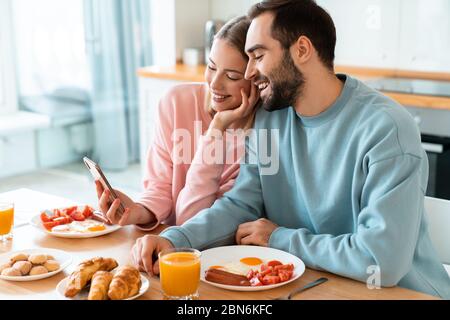 Portrait of young joyful couple using cellphones and laughing while having breakfast in cozy kitchen at home Stock Photo