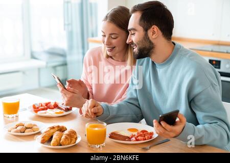 Portrait of young joyful couple using cellphones and smiling while having breakfast in cozy kitchen at home Stock Photo