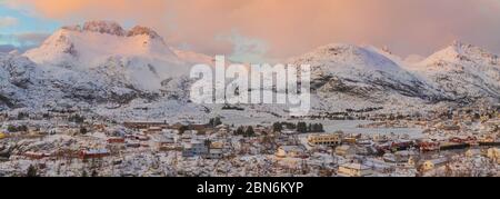 Panromaic view at snow covered mountains. Sunset, orange light on snow. Fishing village at foot of the mountains with traditionar norwich houses