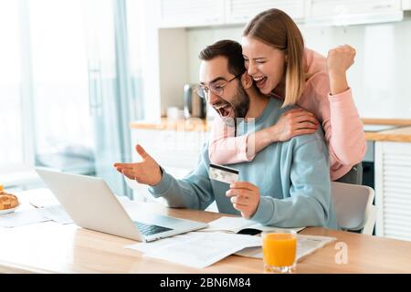 Portrait of young excited couple hugging while working with laptop and holding credit card in cozy kitchen at home Stock Photo