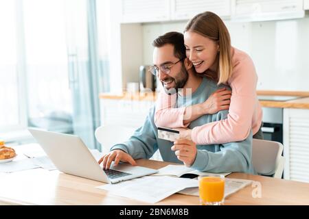 Portrait of young joyful couple hugging while working with laptop and holding credit card in cozy kitchen at home Stock Photo