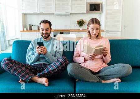 Portrait of young nice couple using cellphone and reading book while sitting on sofa in cozy room at home Stock Photo