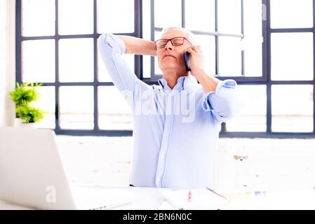 Shot of careworn senior businessman with hand on head talking with somebody while sitting behind his laptop in the office. Stock Photo