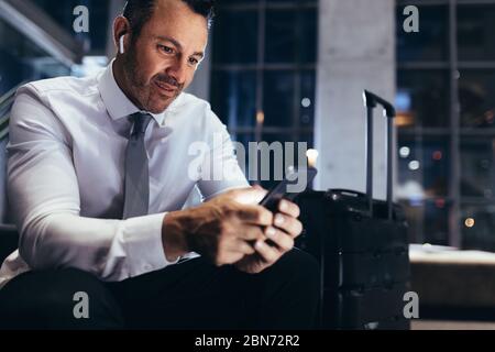 Businessman waiting at the airport first class lounge with luggage next to him. Man in formalwear sitting at airport departure area using his smart ph Stock Photo