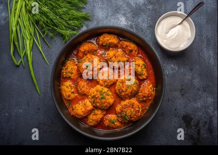 Homemade meatballs in tomato sauce in a saucepan against a dark background. Next to the saucer with sour cream and greens Stock Photo