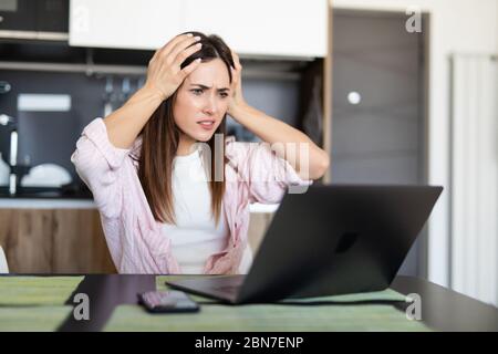 Shocked cute young pretty woman standing in kitchen indoors at home using laptop computer Stock Photo