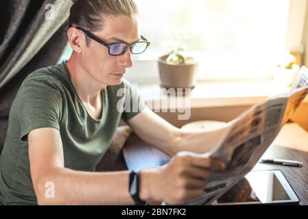 A young handsome man reading interesting hot news articles in a newspaper and drinking Cup of cappuccino coffee near the window. Concept of spending t Stock Photo