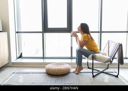 Young woman sitting on modern chair in front of window relaxing in her living room and drinking coffee or tea Stock Photo